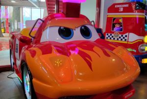 A children’s amusement ride designed as a bright orange and red race car with a screen showing a child's image sits near another ride resembling a fire truck. Set within an indoor water park, this vibrant scene is part of an exciting Idaho theme park adventure.