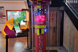 Image of a commercial boxing arcade machine labeled "Boxer Glove" with a red punching bag and electronic display. It is located next to a coin-operated kiddie ride inside an indoor water park entertainment venue.