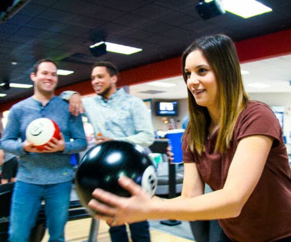 Young woman bowling a ball with two friends standing in the background watching and smiling
