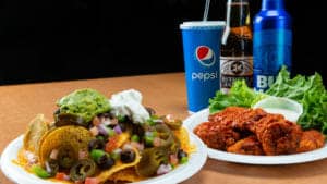 A plate of nachos and a plate of wings next to a soda and beer