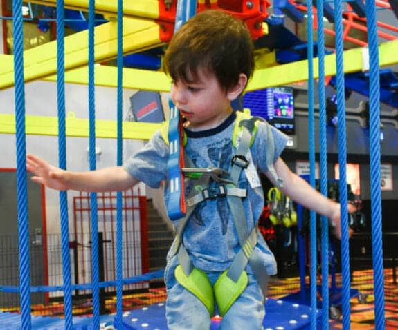 A young child wearing a harness explores a ropes course in an indoor play area, surrounded by the excitement of an indoor water park.