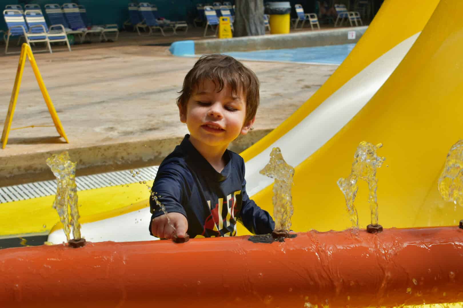Young child playing with water jets at a splash pad, smiling with eyes closed, wearing a long-sleeved shirt. Yellow slide and empty lounge chairs are visible in the background of this Idaho theme park.