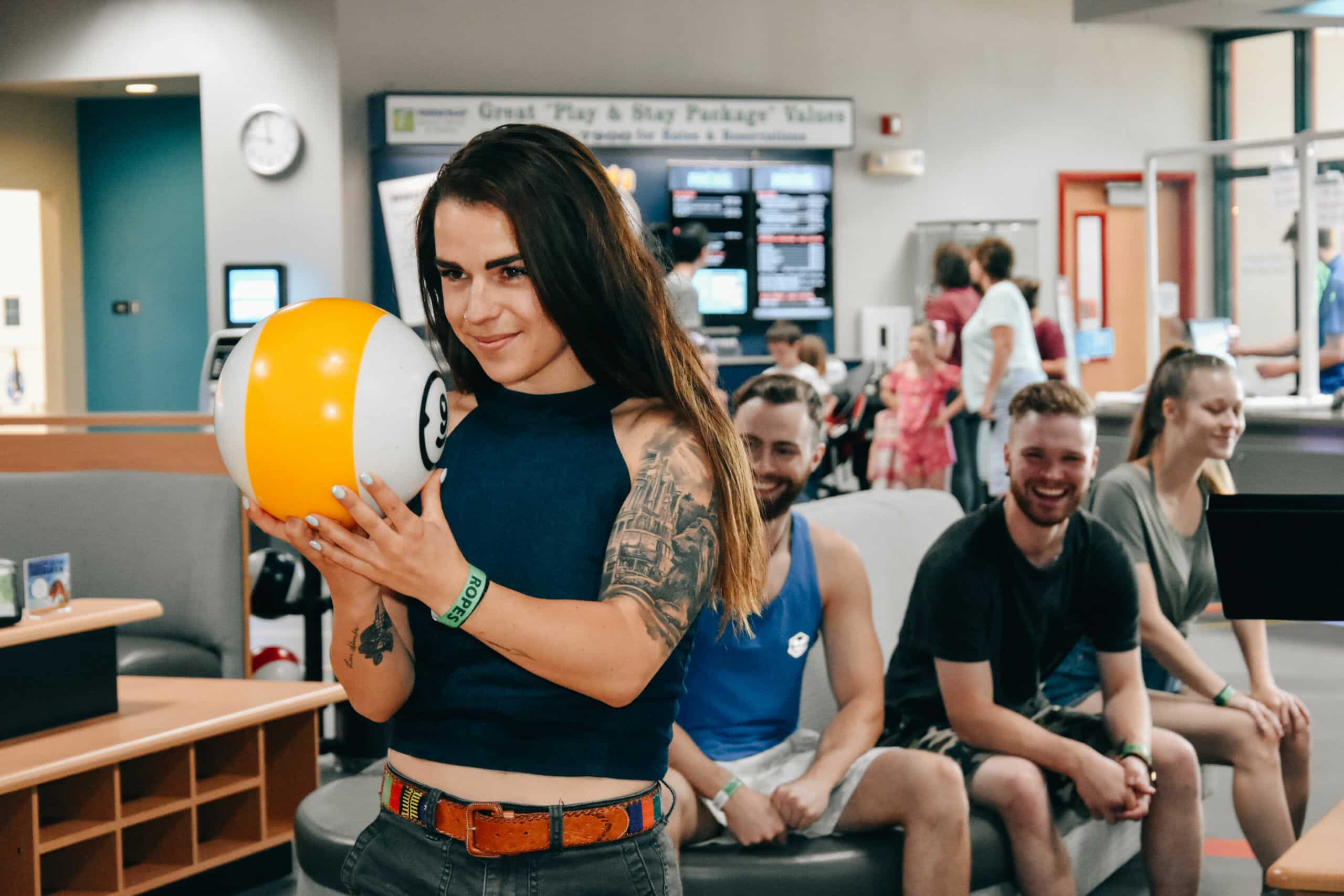A woman holding a yellow and white bowling ball stands in front of a group of people sitting on a bench, some of whom are smiling and talking. The setting appears to be an amusement park in Idaho, perhaps with activities like laser tag Cour d'Alene nearby.