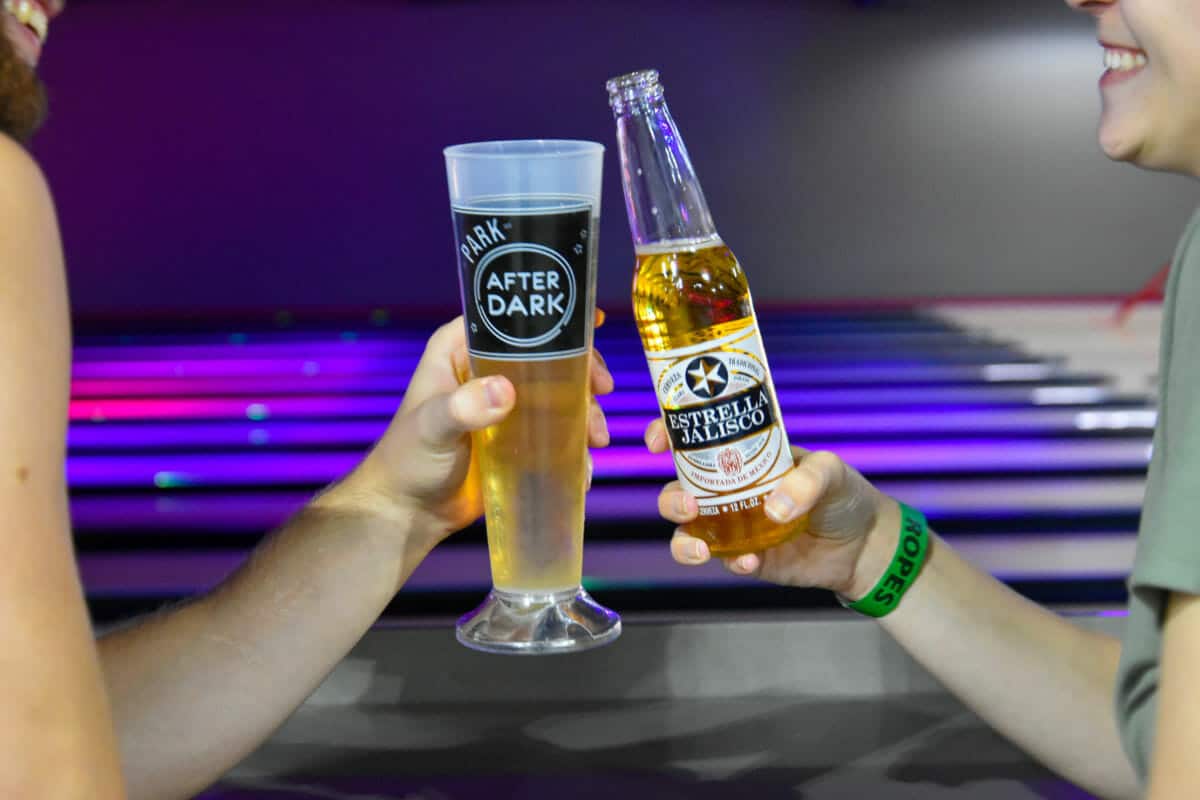 Two people clinking a beer glass and a bottle of Estrella Jalisco beer, with a bowling alley and the excitement of an arcade in Idaho in the background.