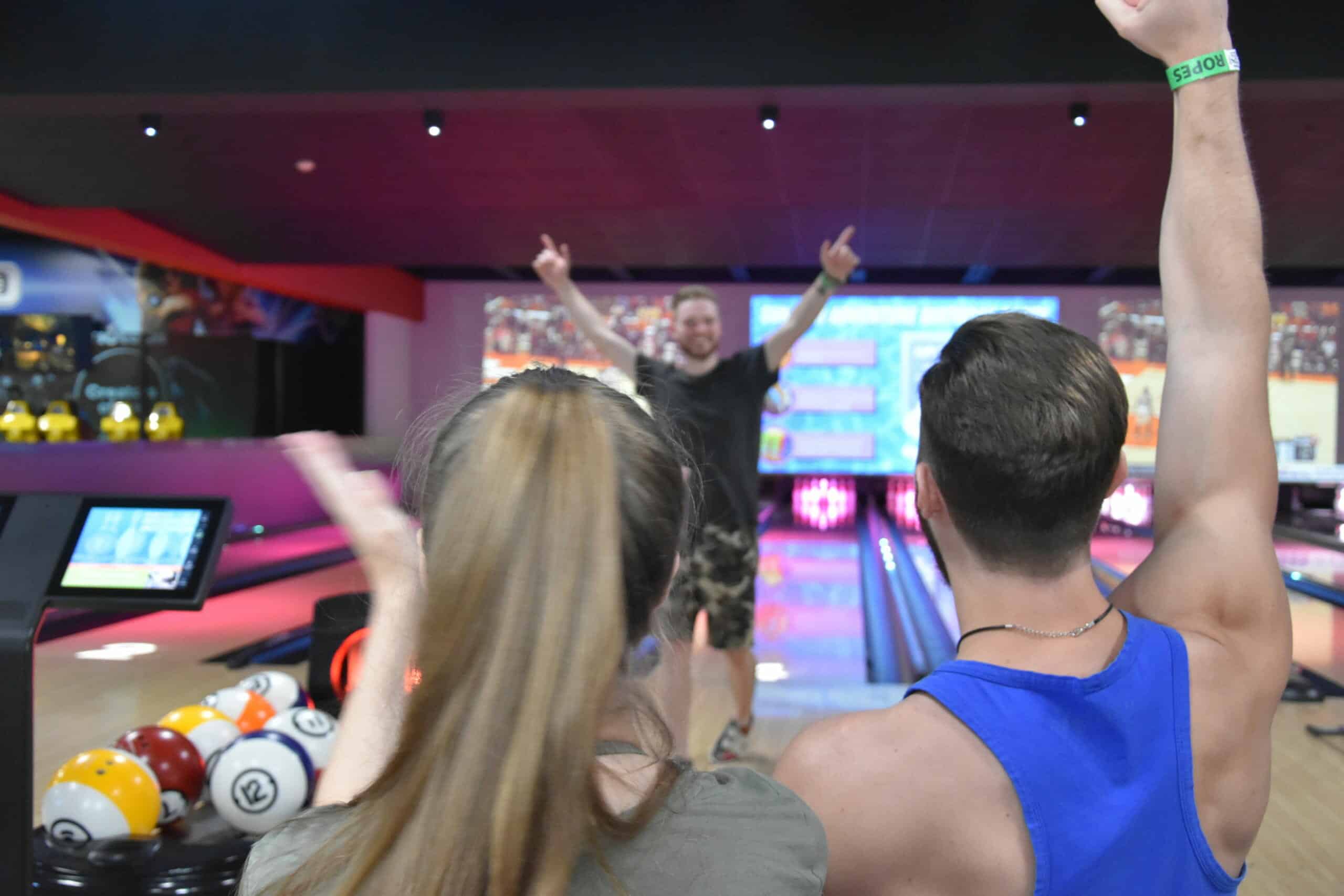Two people celebrate as a man wearing a camo outfit raises his arms in victory at a bowling alley. Sporting equipment and a screen are visible in the background, reminiscent of the joyous moments often found at an arcade in Idaho.