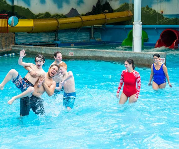 Adults and children enjoy a lively game in an indoor water park pool, with slides and painted scenery in the background at an Idaho theme park.