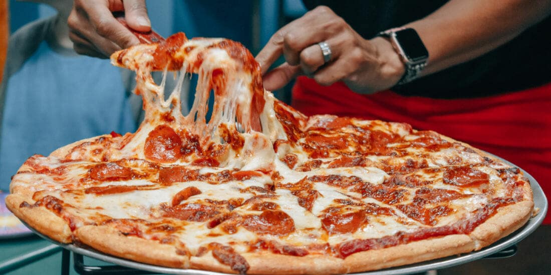 A person lifts a cheesy slice from a pepperoni pizza on a metal stand, perhaps fueling up before an adventure at the nearby amusement park in Idaho. Another individual is partially visible in the background.
