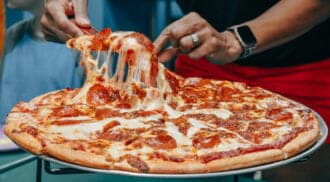 A person lifts a cheesy slice from a pepperoni pizza on a metal stand, perhaps fueling up before an adventure at the nearby amusement park in Idaho. Another individual is partially visible in the background.