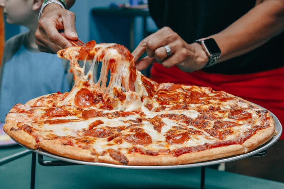 A person lifts a cheesy slice from a pepperoni pizza on a metal stand, perhaps fueling up before an adventure at the nearby amusement park in Idaho. Another individual is partially visible in the background.