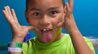 A child wearing a bright green "Triple Play Family Fun Park" shirt playfully makes a funny face by pulling at his cheeks and sticking out his tongue, embodying the carefree spirit of an amusement park in Idaho.
