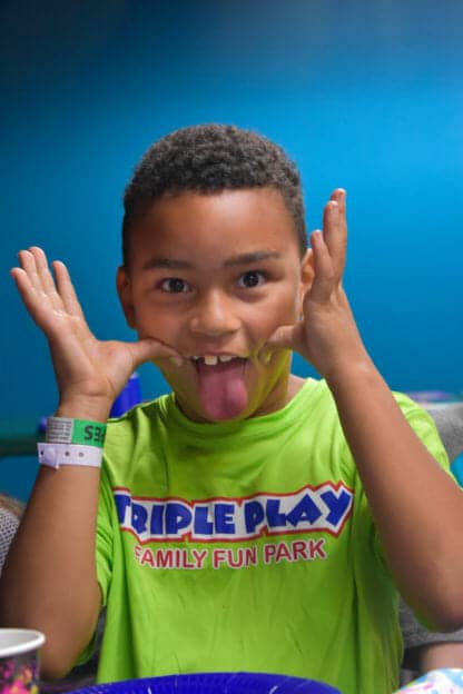 A child wearing a bright green "Triple Play Family Fun Park" shirt playfully makes a funny face by pulling at his cheeks and sticking out his tongue, embodying the carefree spirit of an amusement park in Idaho.
