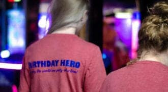 Two individuals with long hair and wearing red T-shirts with "BIRTHDAY HERO" printed on the back stand in a dimly lit room at an arcade in Idaho, with colorful lights in the background.