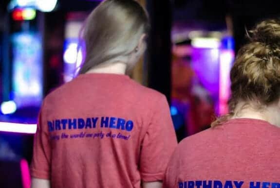 Two individuals with long hair and wearing red T-shirts with "BIRTHDAY HERO" printed on the back stand in a dimly lit room at an arcade in Idaho, with colorful lights in the background.