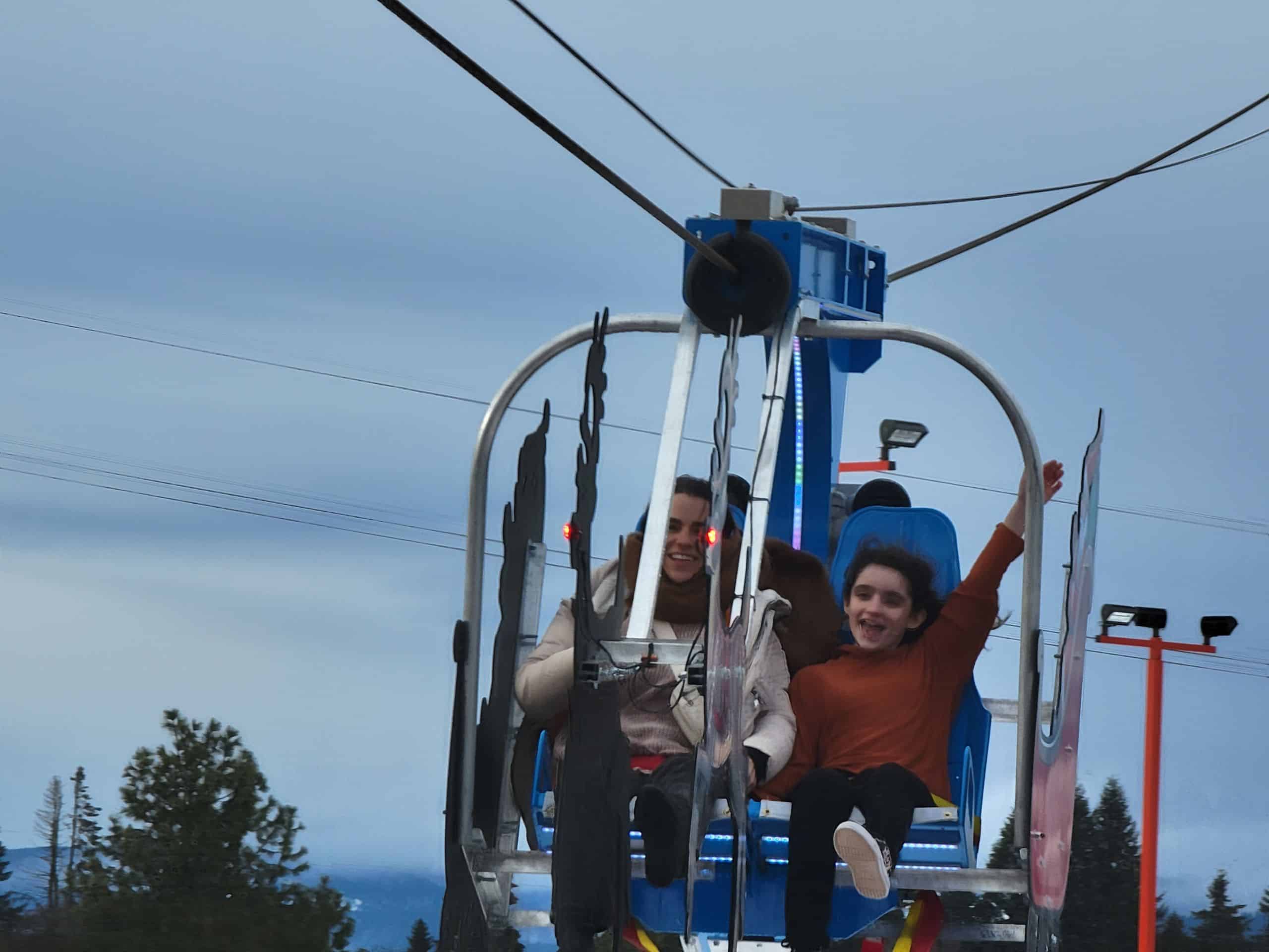 Two people, seated in an amusement ride with harnesses, appear to be enjoying the experience. The background shows a cloudy sky and trees, reminiscent of the outdoor thrills you'd find at an arcade in Idaho.