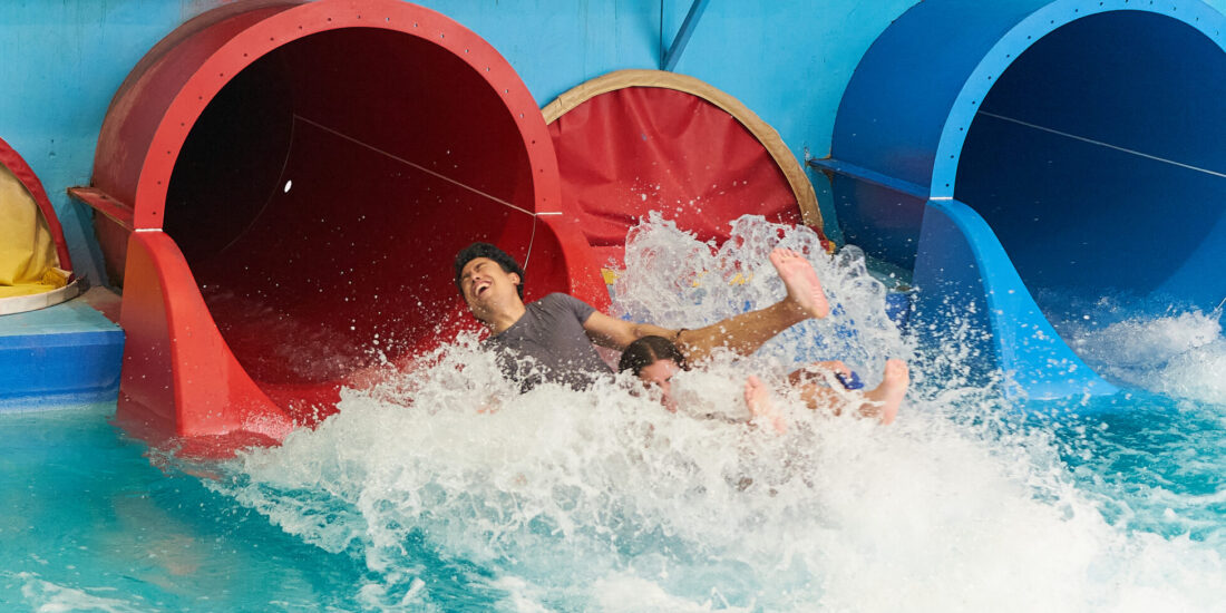 Two people laughing as they splash into a pool at the bottom of a large red indoor water slide.