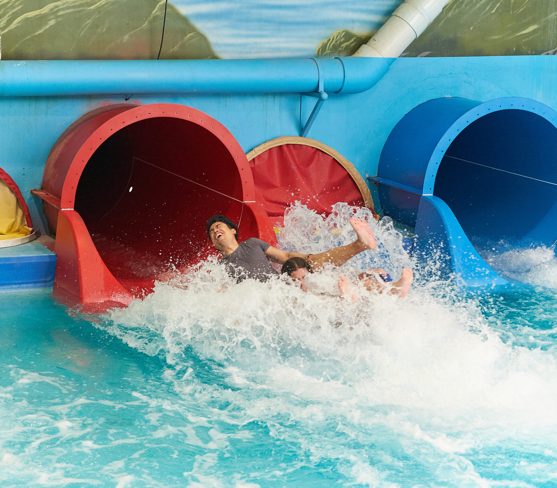 Two people laughing as they splash into a pool at the bottom of a large red indoor water slide.