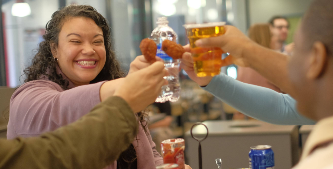 A smiling woman and friends clinking chicken wings, a water bottle, and a beer together over a meal at a restaurant table.