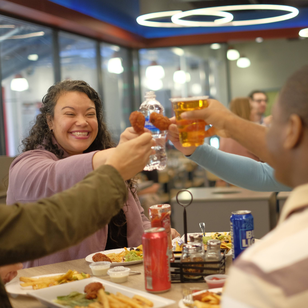 A smiling woman and friends clinking chicken wings, a water bottle, and a beer together over a meal at a restaurant table.