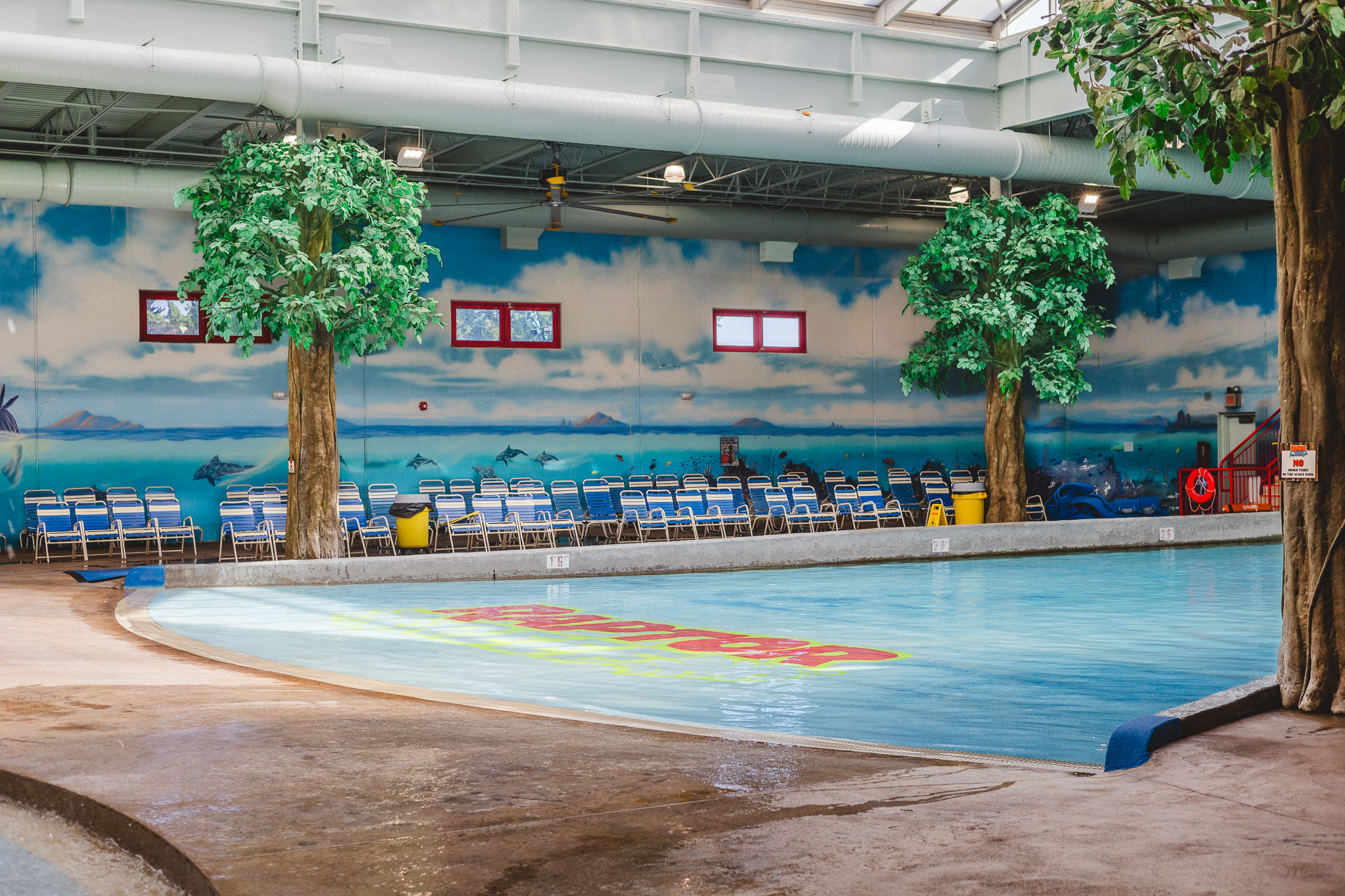 Zero-depth entry wave pool at Raptor Reef indoor water park lined with artificial trees, lounge chairs, and an ocean-themed wall mural.