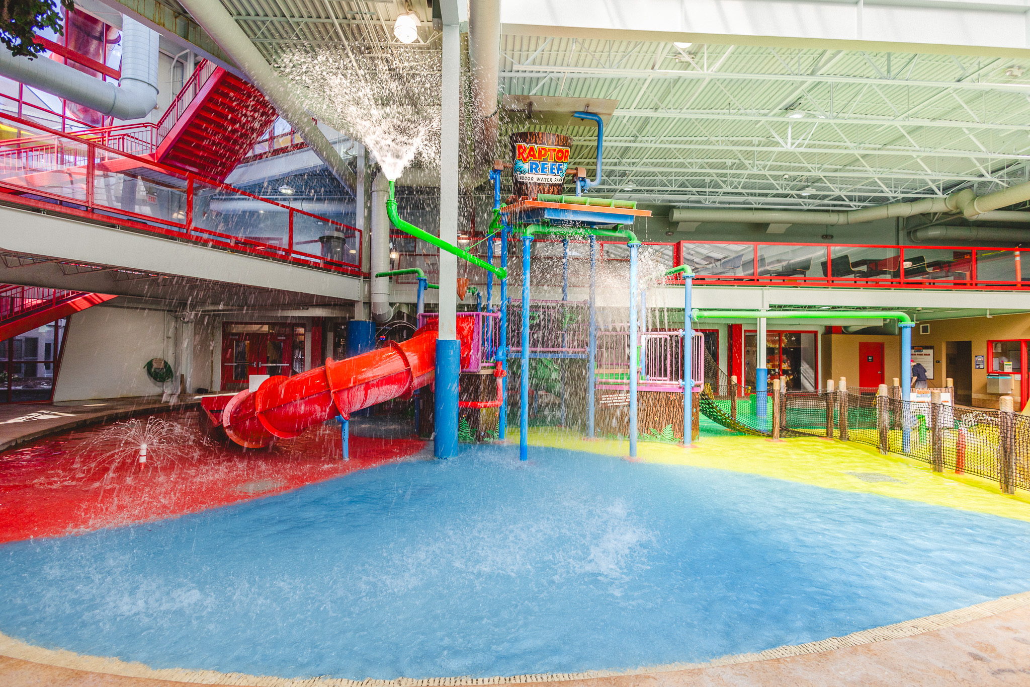 An indoor water park play structure at Raptor Reef featuring a red tube slide, splash fountains, climbing platforms, and a tipping water bucket over colorful blue, yellow, and red shallow pool flooring.